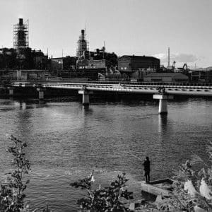 Photo of serene river with bridge and industrial buildings backdrop.