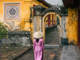 Woman in traditional Vietnamese attire walking towards historic yellow archways in Historic Old Imperial Citadel of Hue .