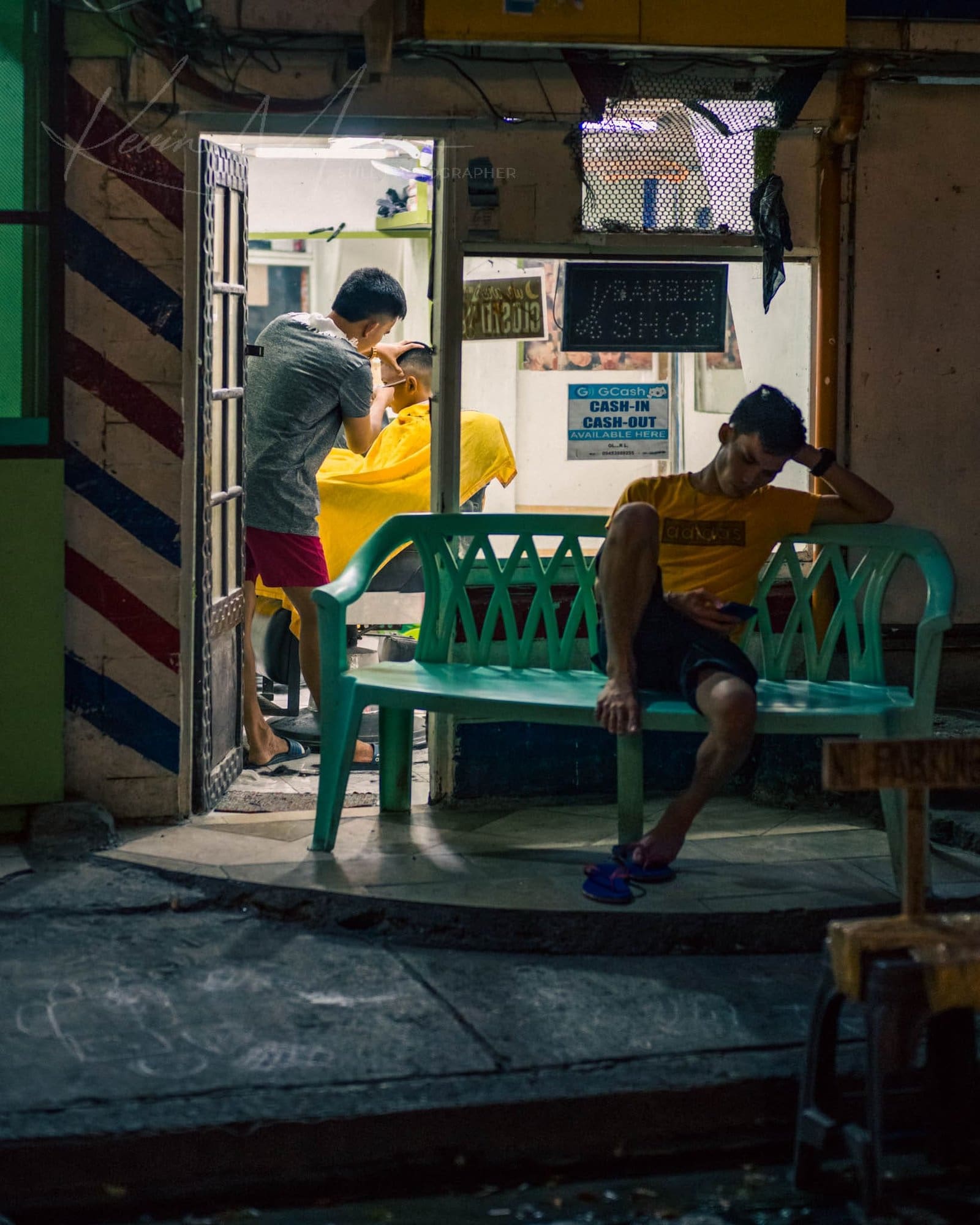 Barber giving haircut in busy nighttime barbershop scene.