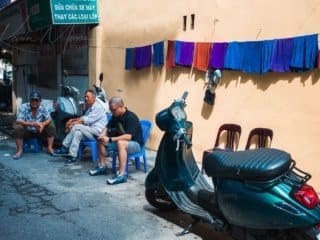 Hanoi Alleyway Life: Local Chats and Blue Scooter Scene Casual street chatter in a narrow Hanoi alleyway with a blue scooter and colorful hanging towels.
