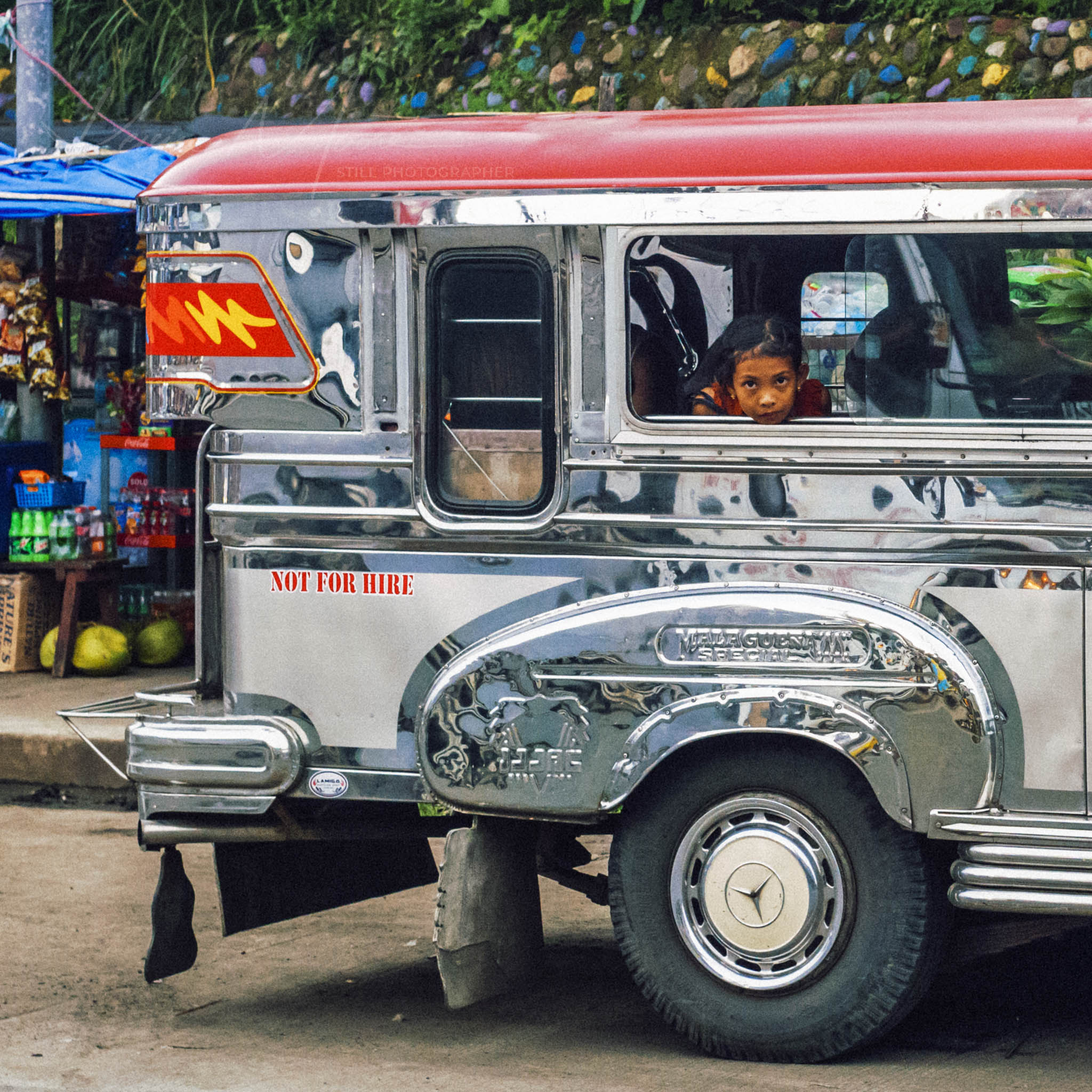 Child peeks from decorated Jeepney at a vibrant Filipino market.