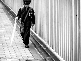 Visually impaired elderly man confidently walks by fence with walking stick and camera.