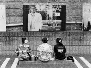 Three individuals in kimonos under a billboard of a man in Western attire, Japan.
