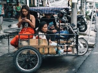 Women using smartphone in a cargo tricycle on a busy urban street after rain.