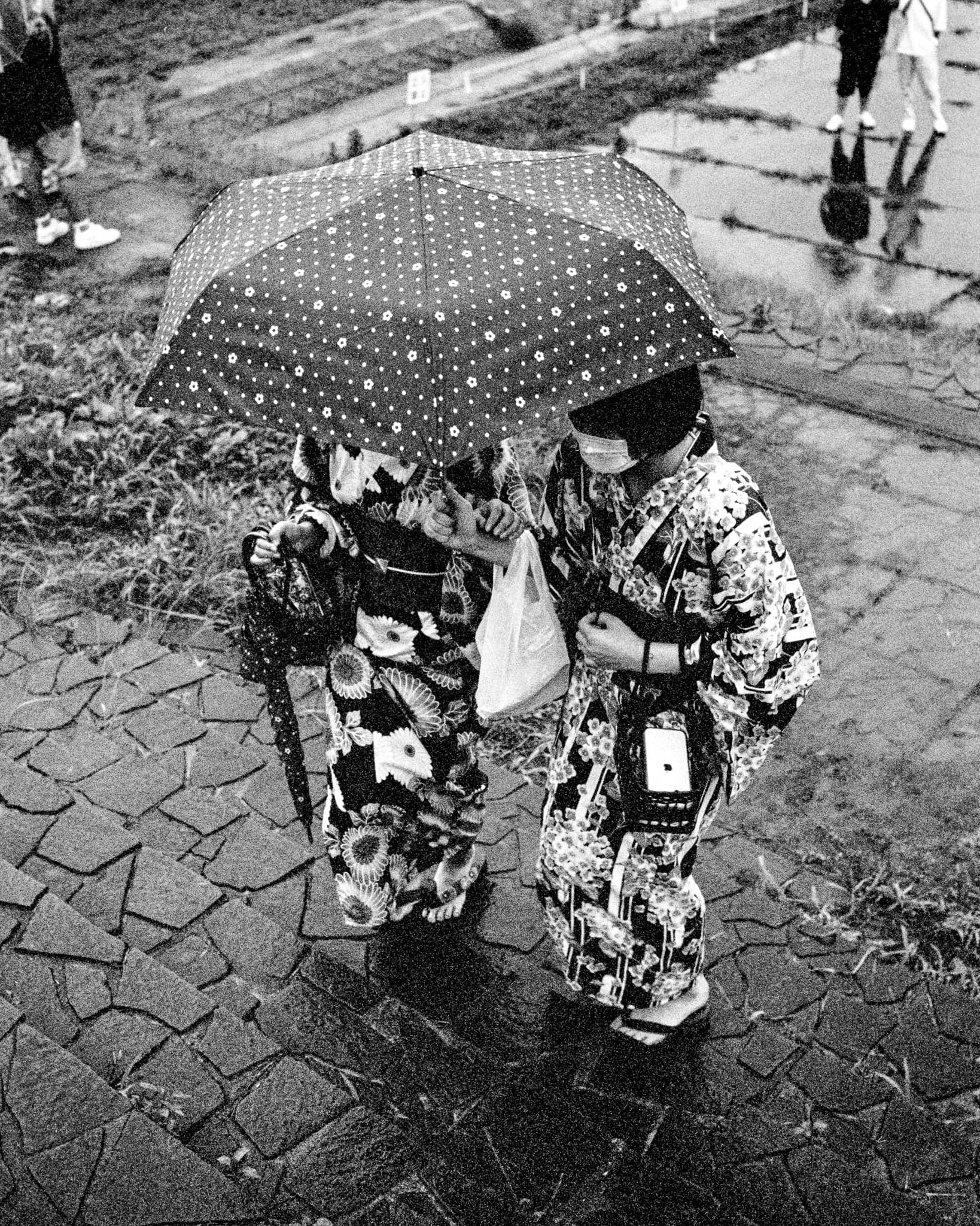 B&W film photo image, taken in Iwaki City, Fukushima, Japan, captures two young women clad in traditional Japanese yukata huddled under a shared umbrella