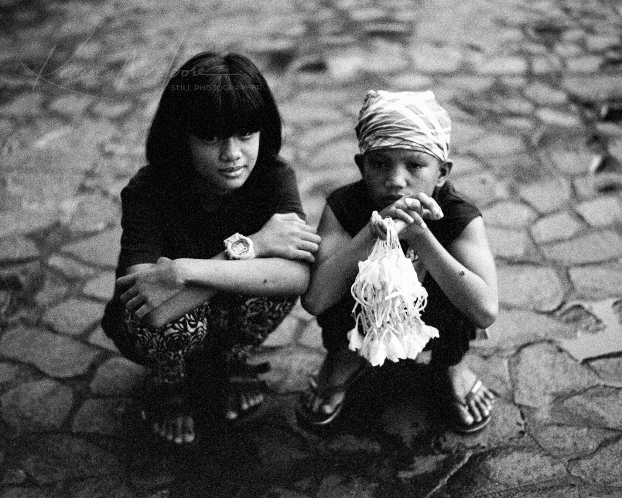 Filipino children selling Sampaguita flowers on cobblestone in moody black and white portrait