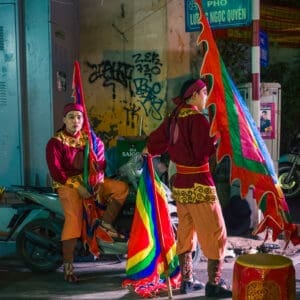 Street performers in vibrant costumes under spotlight on Hanois graffiti-covered city street at night.