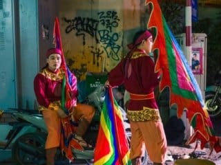 Hanoi Night: Spotlight on Heritage Street Performers Street performers in vibrant costumes under spotlight on Hanois graffiti-covered city street at night.