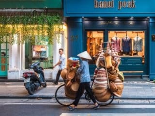 Vietnamese vendor transporting goods on a bicycle amid vibrant cityscape in Hanoi.