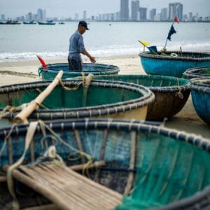 Fisherman with traditional Vietnamese Thung Chai boats on Da Nang beach, modern cityscape in background.