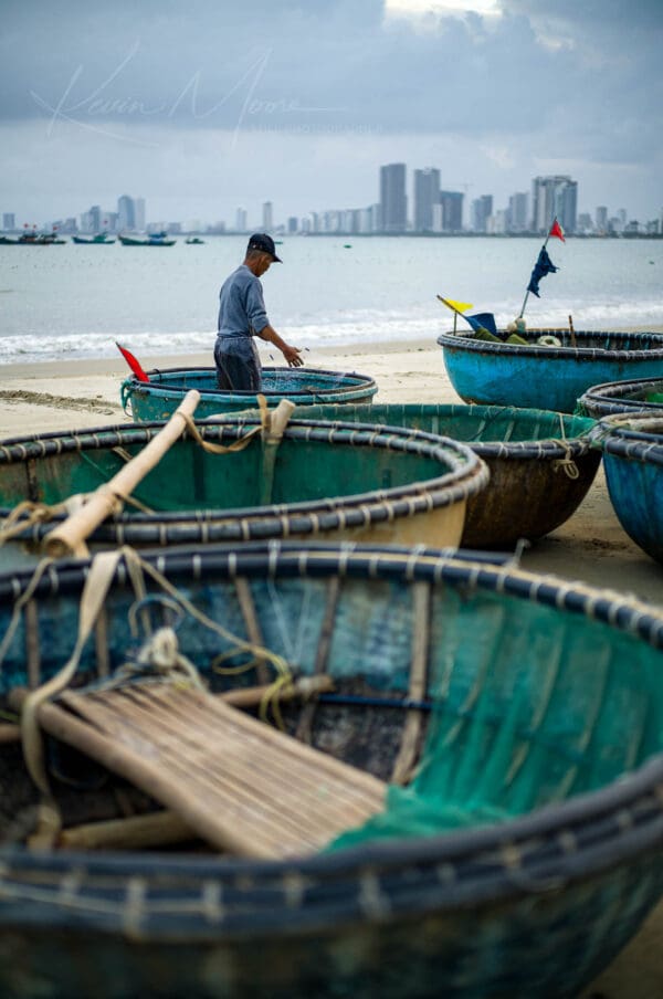 Fisherman with traditional Vietnamese Thung Chai boats on Da Nang beach, modern cityscape in background.