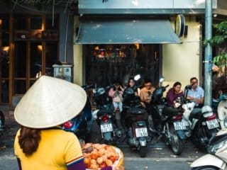 Woman selling fresh persimmons on a bustling Hanoi Vietnam street with coffee shop and motorbikes.