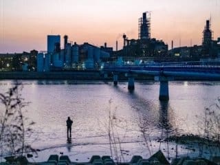 Solitary figure fishing by a serene inlet against a twilight industrial cityscape.