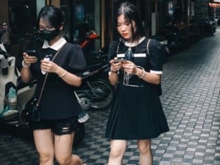 Tech-Engrossed Vietnamese Girls Stroll Tranquil Hanoi Street Two young Vietnamese women engrossed in smartphones on a tiled pedestrian street in Southeast Asia.