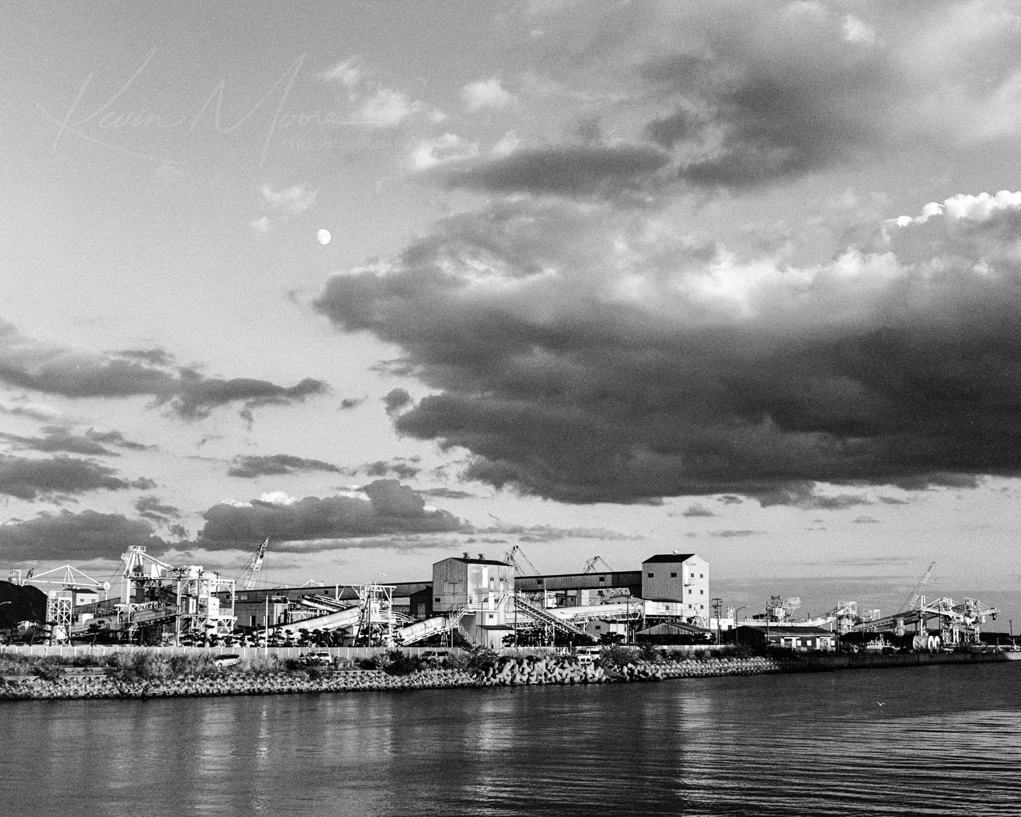 Industrial riverfront with factories, cranes, and reflective water under a cloudy sky with distant moon.