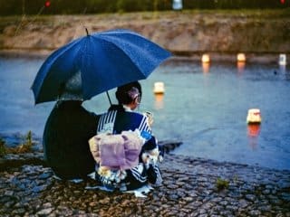 A couple in traditional Japanese attire sitting by a river with floating lanterns at twilight.