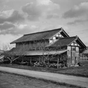 Weathered Japanese barn raised on posts under a dramatic winter sky, bare trees in front, rural Iwaki, Fukushima