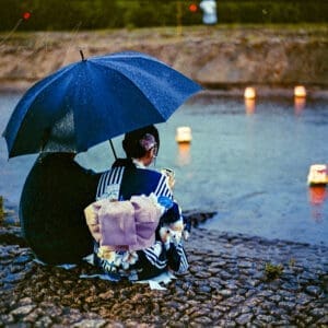 A couple in traditional Japanese attire sitting by a river with floating lanterns at twilight.