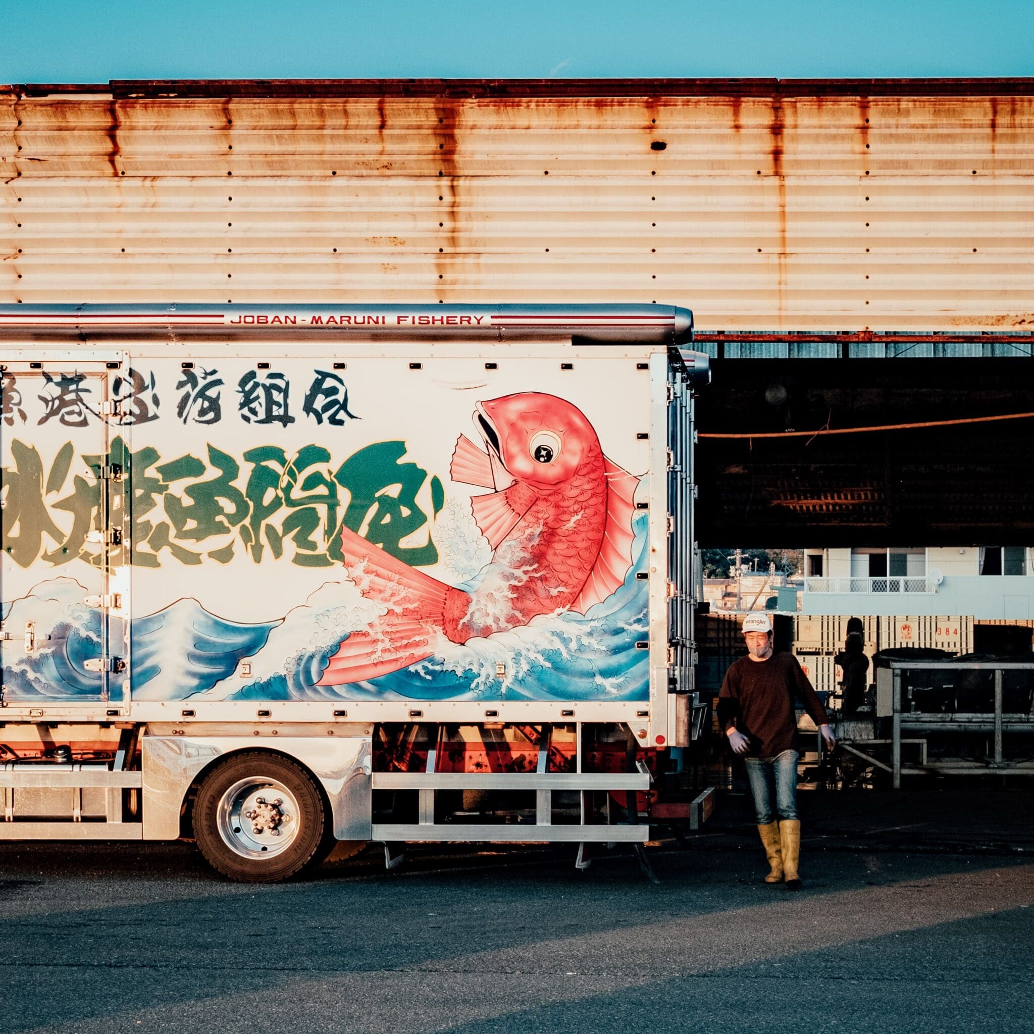 Worker preparing seafood delivery in traditional Japanese-art adorned Joban-Maruni Fishery truck at sunset.