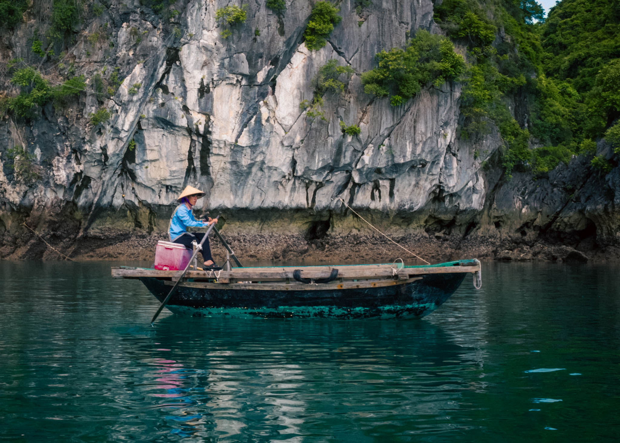 Solitary woman rowing a rustic boat in serene Ha Long Bay emerald waters against a limestone cliff backdrop.