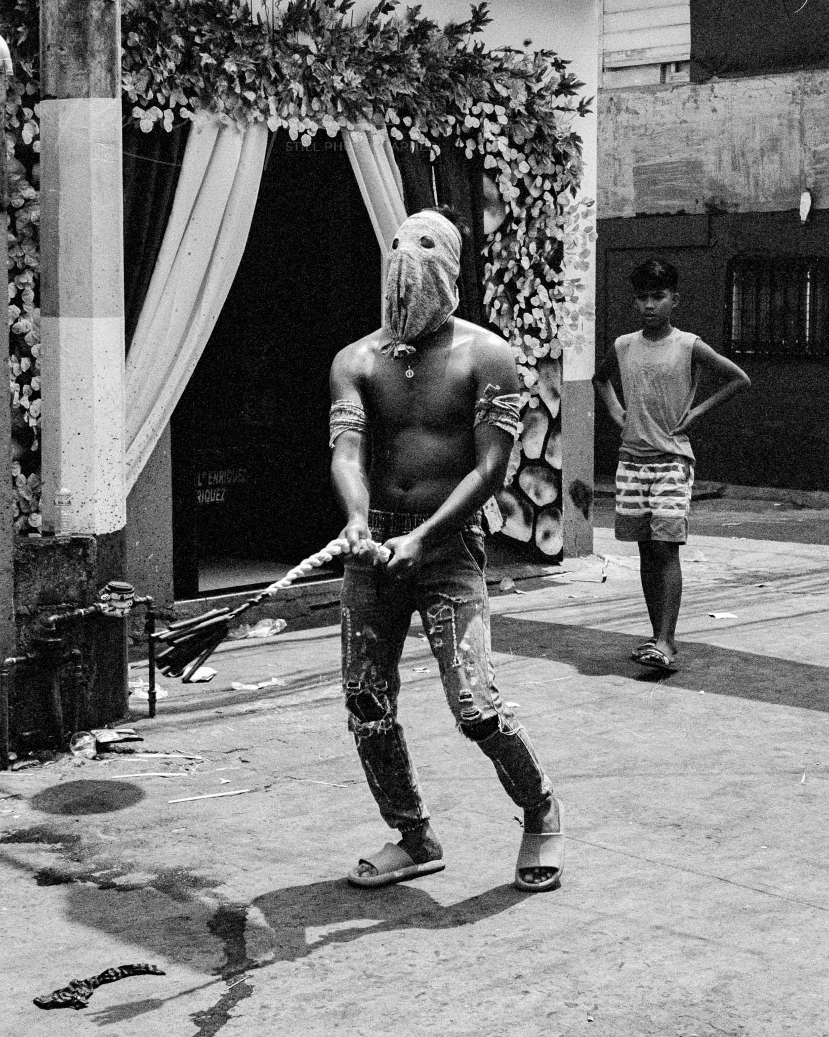 Shirtless man in bird mask with staff on vibrant urban street.