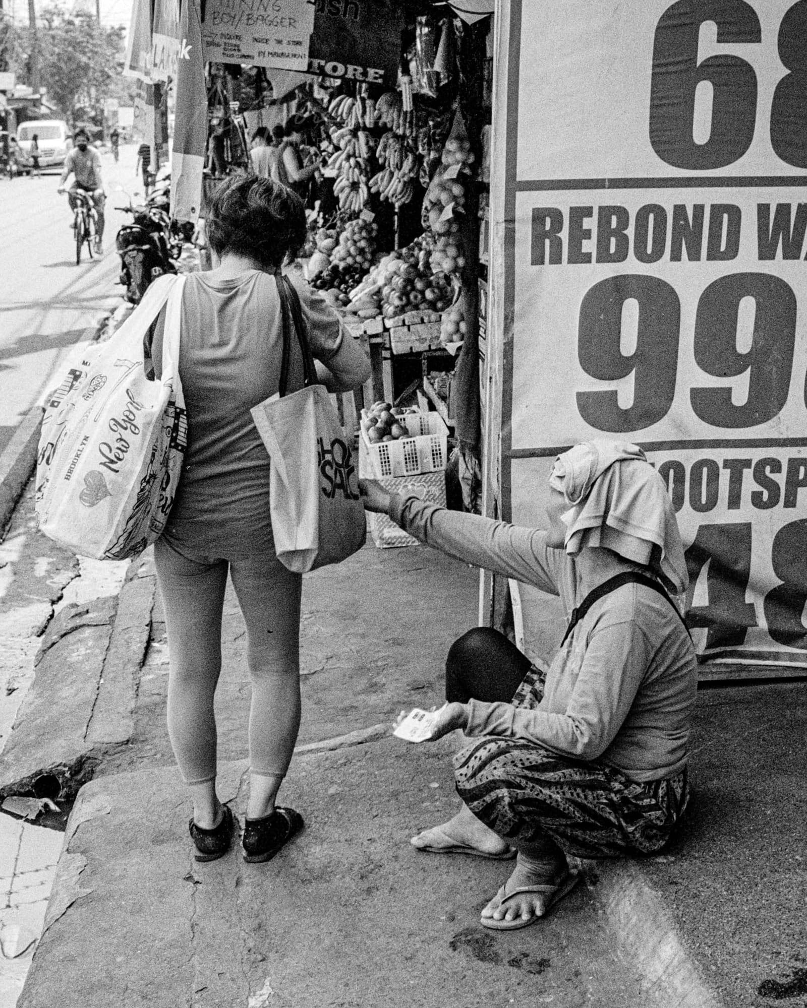 Woman giving money to seated person by fruit store in busy urban street.