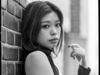 Young woman in casual dress leans against brick wall in evocative black and white portrait.
