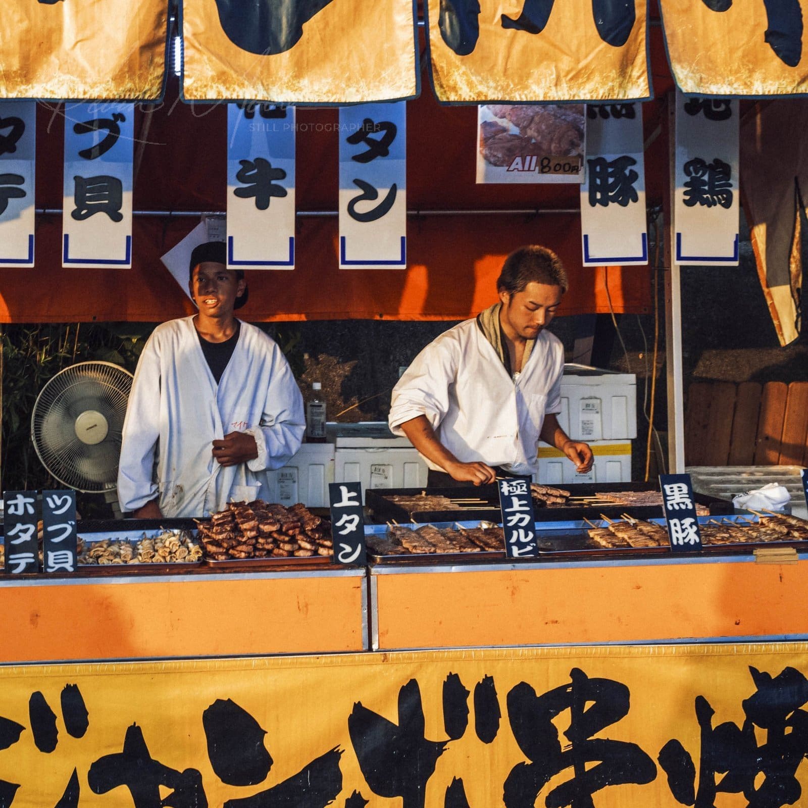 Japanese festival food stall with vendors grilling skewered meats at dusk.