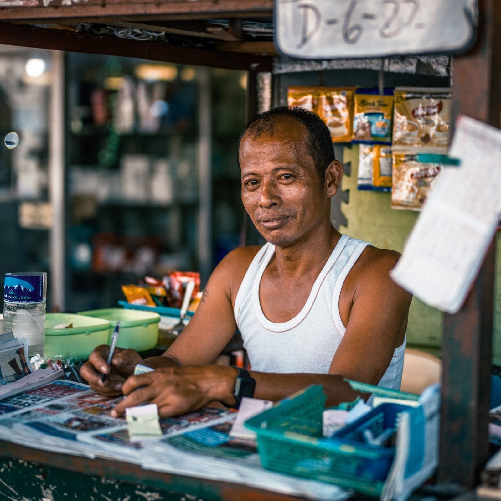 Vendor at tropical market stall surrounded by snacks and goods.